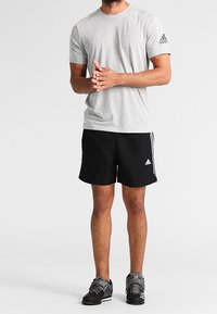 Maglietta sportiva grigia a maniche corte con logo Adidas nero sulla spalla sinistra, abbinata a pantaloncini neri con strisce bianche e scarpe da ginnastica nere.