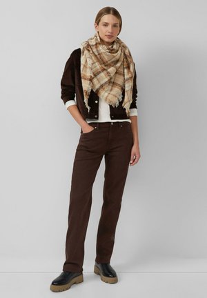 Frau steht mit den Händen in den Taschen, trägt eine braune Jacke, ein weißes Hemd, braune Hose, einen beigen karierten Schal und schwarze Stiefel mit beigen Sohlen.