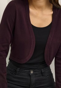 Cardigan corto bordeaux con una texture liscia, tessuto morbido, design a apertura frontale e maniche lunghe, indossato sopra una maglietta nera.