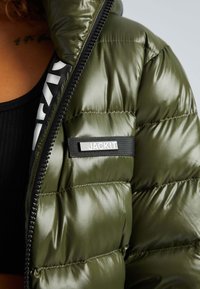 Olijfgroene pufferjack met glanzende afwerking, met horizontale stiksels, een ritsluiting en een merketiket op de linkerborst.