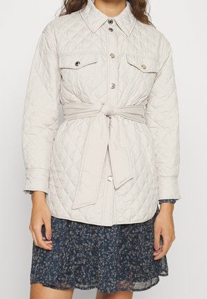 Femme portant une veste matelassée beige clair avec des boutons-pression, des poches poitrine et une ceinture en tissu nouée sur une robe fleurie foncée.