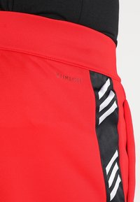 Gros plan sur un pantalon de sport rouge avec un panneau latéral noir, comportant trois rayures blanches diagonales et le texte "CLIMALITE" sur la ceinture.