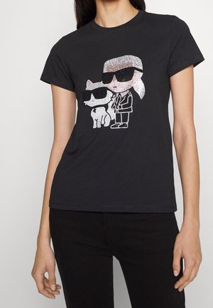 Femme portant un t-shirt noir avec un dessin cartoon en strass représentant une personne et un chien, tous deux avec des lunettes de soleil.