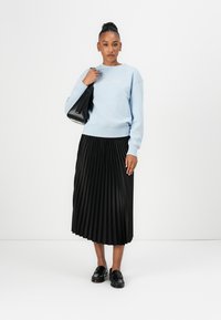 Light blue sweatshirt with white text, black pleated midi skirt, and black loafers. Model holds a black tote bag. Simple design and solid colors.
