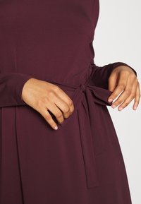 Robe bordeaux à manches longues avec une ceinture à la taille. Le tissu est lisse avec un éclat subtil. Deux poches latérales sont visibles.