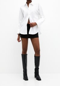 Chemise blanche à boutons, manches longues, mini-jupe noire et bottes noires à talons montant jusqu'aux genoux. Tissu lisse, design structuré et lignes épurées.