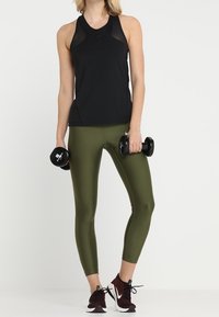 Czarny bezrękawnik sportowy, oliwkowo-zielone dopasowane legginsy i czerwone buty sportowe. Osoba trzyma czarne hantle.