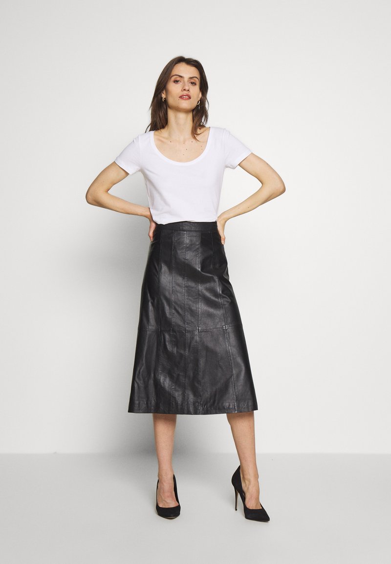 Falda midi de cuero negro con cintura alta y diseño de paneles, combinada con una blusa blanca de manga corta y tacones negros de punta.