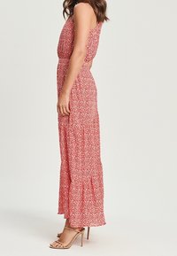 Robe maxi florale rouge avec un corsage ajusté et une jupe à volants. Elle présente des fines bretelles et une texture fluide. Associée à des sandales à talons beiges.
