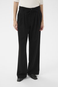 Schwarze, weit geschnittene Hose mit hoher Taille, Falten, Gürtelschlaufen und gebundenem Bund, kombiniert mit spitzen schwarzen Schuhen. Glatte Stoffstruktur.