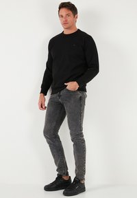 Fekete hosszú ujjú pulóver kerek nyakkivágással, szürke slim-fit farmerrel és fekete sportcipővel viselve. Sima anyag, minimalista dizájn.