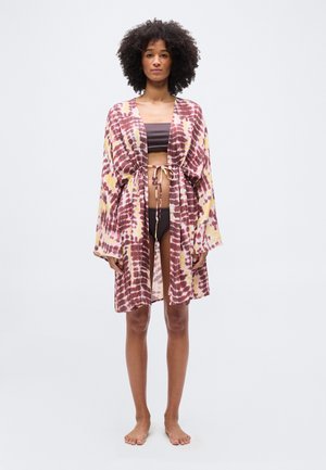 Kvinde med krøllet hår iført en brun bikini og en løs, tie-dye mønstret kjole i pink, gul og brun, bundet om livet.