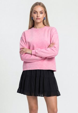Ung kvinde med langt blondt hår, krydsede arme, iført pink sweatshirt, sort lagdelt nederdel og blå stjerneformede øreringe, på ensfarvet baggrund.