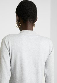 Maglione grigio a maglia con colletto a coste, maniche lunghe e una texture liscia. Sono visibili orecchini a cerchio dorati nell'orecchio.
