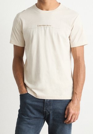 Mężczyzna w beżowej koszulce z krótkim rękawem Calvin Klein Jeans i niebieskich dżinsach, stojący na białym tle z jedną ręką w kieszeni.