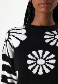 Black sweater featuring large white floral patterns, ribbed neckline, and textured knit fabric. Suitable for casual wear.