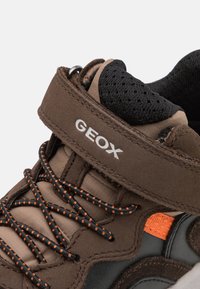Zapato de tela marrón con una correa de Velcro, forro de malla negra, acento naranja y cordones negros. Marca GEox destacada en la correa.
