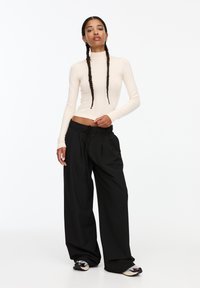 Cremfarbener Rollkragenpullover kombiniert mit schwarzen weit geschnittenen Hosen. Die Hosen haben eine Kordel zum Verstellen und Faltendetails. Model trägt gemusterte Schuhe.