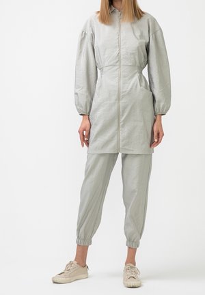 Femme porte une longue veste zippée gris clair avec taille froncée, pantalon de jogging assorti et baskets beiges, debout sur un fond blanc.