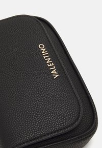 Bolso de cuero negro texturizado con forma redondeada, que presenta un logotipo dorado "Valentino" en la parte delantera y un cierre de cremallera suave.