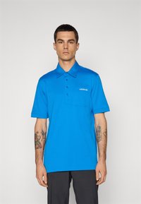 Blå polo-skjorte med knapplukking, korte ærmer og brystlomme. Har adidas-logo brodert i hvitt. Myk, glatt tekstur.
