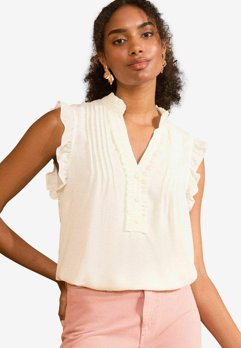 Blusa senza maniche color crema con colletto arricciato e chiusura a bottoni, caratterizzata da pieghe verticali e una texture morbida e liscia. Abbinata a pantaloni rosa.