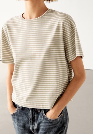 Personne portant une chemise à manches courtes beige et blanche à rayures, les mains dans les poches d'un jean bleu taille haute, devant un fond uni.