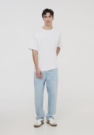 T-shirt bianco a maniche corte abbinato a jeans relaxed-fit azzurri e sneakers bianche con dettagli neri. Sfondo semplice, design minimalista.