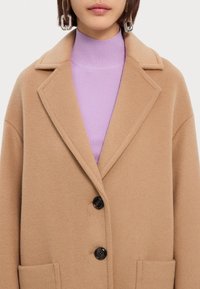 Abrigo de lana beige con solapa, cierre de dos botones y bolsillos delanteros; superpuesto a un cuello alto de canalé morado.