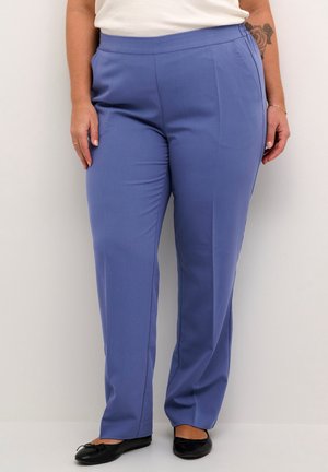 Vrouw draagt rechte blauwe broek met zijzakken, een wit topje, zwarte balletschoenen en een zichtbare rozentatoeage op haar arm.