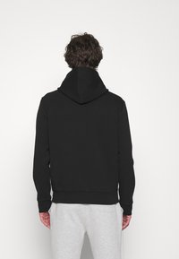 Felpa nera con zip e cappuccio grande, con polsini e fascia in costina, vista da dietro, indossata sopra pantaloni sportivi grigi. Tessuto dalla texture liscia.