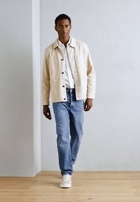 Giacca beige chiaro con bottoni, camicia bianca sotto, jeans blu e sneakers bianche. Il modello è in piedi contro uno sfondo neutro.