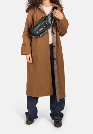 Persoon draagt een lange bruine jas, blauwe spijkerbroek, zwarte schoenen en een groene gewatteerde crossbodytas met een gevlochten riem.