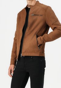 Uomo che indossa una giacca in suede marrone con cerniera sul petto e tasche laterali, camicia nera e jeans neri, con una mano nella tasca della giacca.