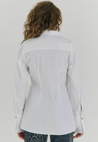 Hvid skjorte med knapper, der har en rygpanel, lange ærmer med sidelæg og sølvfarvede knapdetaljer. Glat tekstur og skræddersyet pasform.