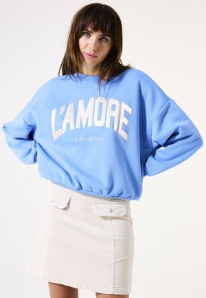 Sweat-shirt bleu clair avec "L'AMORE" imprimé en blanc et beige clair. Associé à une jupe en velours côtelé beige avec des poches à pression.