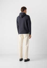 Hanorac cu glugă de culoare bleumarin, din material texturat, cu buzunar tip kangaroo, mansete ribbate, asortat cu pantaloni bej deschis și pantofi negri.