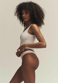 Beige naadloze ondergoed en witte tanktop. De stof lijkt zacht met minimale stiksels. Model poseert zijwaarts tegen een neutrale achtergrond.