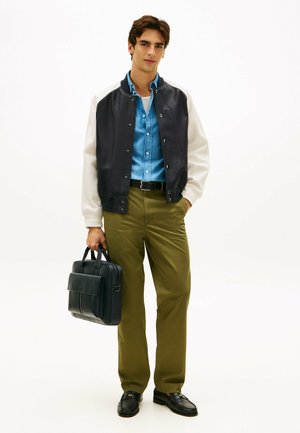 Jonge man in een zwart-wit varsity jas, blauw denim overhemd, olijfgroene broek, die een zwarte leren aktetas vasthoudt tegen een effen achtergrond.