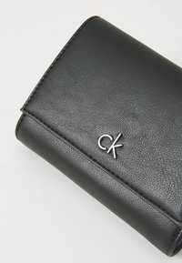 Portefeuille en cuir noir avec une texture grainée, arborant un logo "ck" en argent. Lignes épurées et design minimaliste.