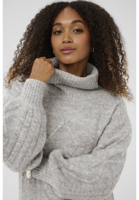 Világosszürke kötött pulóver magas, hajtogatott gallérral és kábelmintás kialakítással. Túlméretezett ujjak és texturált anyag részlet jellemzi.