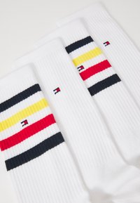 Chaussettes en coton blanc de type crew avec une texture côtelée, présentant des rayures horizontales en marine, rouge, jaune et blanc. Détail de logo discret inclus.