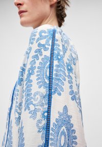 Μπλούζα με μακριά μανίκια από λευκό ύφασμα με περίτεχνα μπλε λουλουδένια σχέδια και λεπτομερή ραφές κατά μήκος των μανικιών. Ελαφριά και υφή.