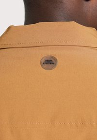 Guleră de cămașă maro cu cusături vizibile și un logo circular etichetat "DENIM ATTITUDE" atașat la spate. Textură de țesătură netedă.