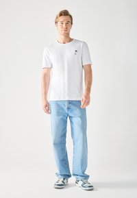Witte t-shirt met korte mouwen en een klein zwart palmbomenontwerp, gecombineerd met lichtblauwe rechte jeans en grijze sneakers.