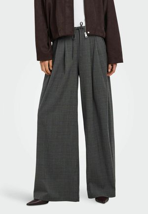 Pantalon large gris à carreaux avec taille à cordon, porté avec des chaussures pointues foncées et une veste marron foncé.