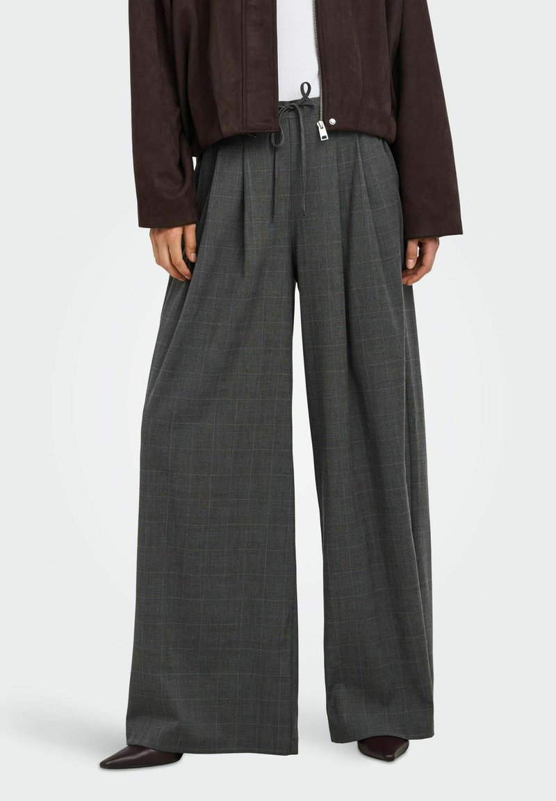Pantalon large gris à carreaux avec taille à cordon, porté avec des chaussures pointues foncées et une veste marron foncé.