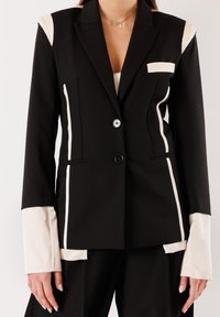 Blazer nero su misura con righe verticali bianche, polsini bianchi che sporgono oltre le maniche e due bottoni neri allacciati sul davanti.