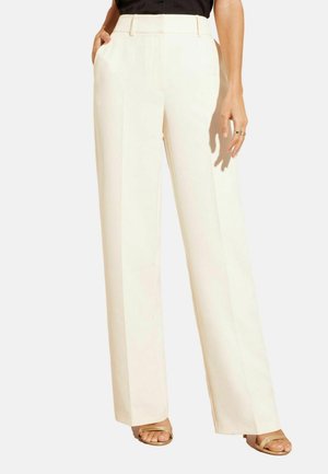 Femme portant un pantalon crème taille haute à jambes droites avec poches latérales et des sandales beige à bout ouvert, debout devant un fond uni.