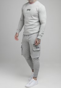 Sudadera gris con cuello redondo y logo en el pecho, combinada con pantalones de chándal grises que tienen solapas en los bolsillos y cremalleras. Zapatillas blancas.